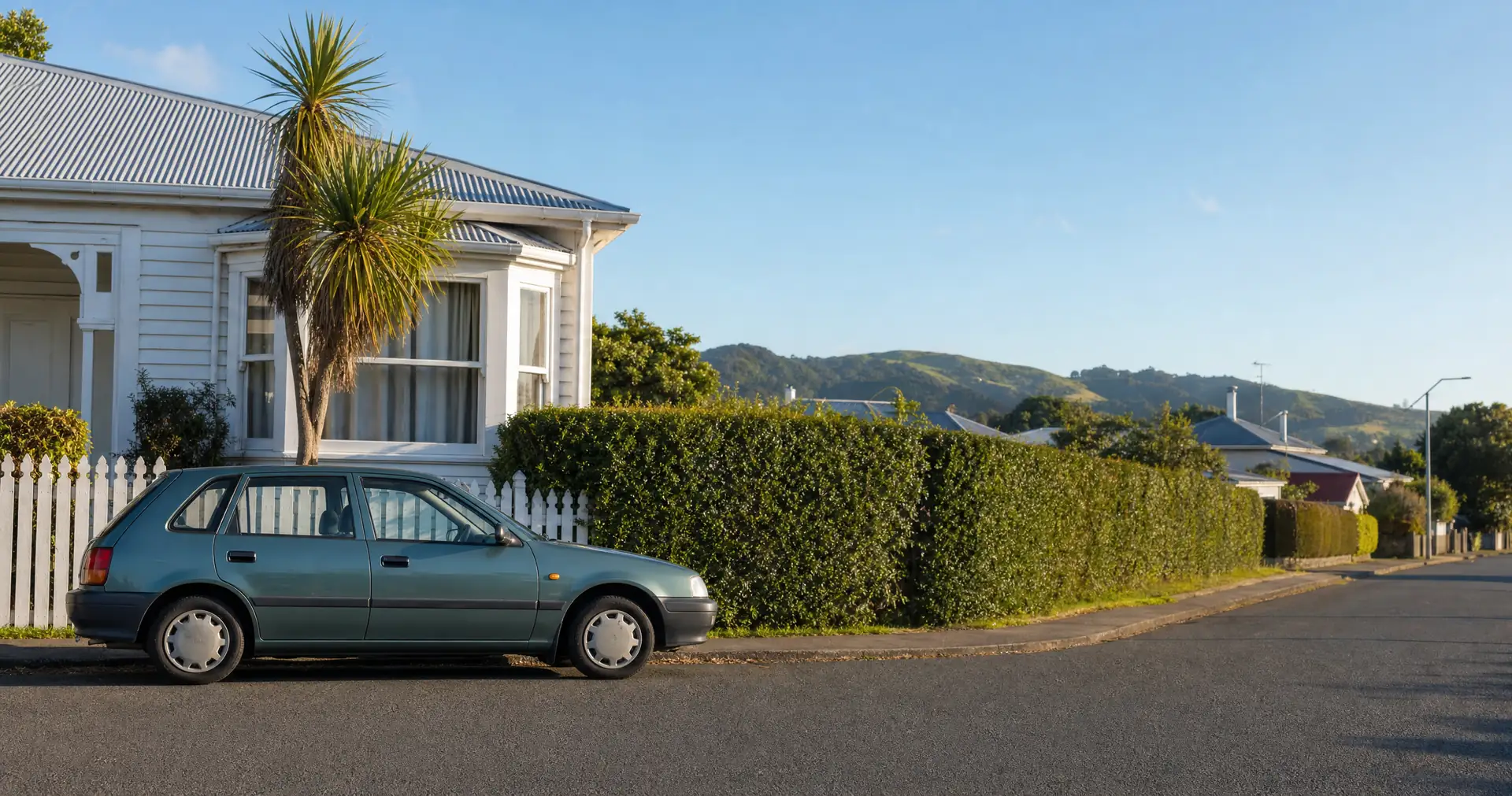 An older small hatchback parked on the street outside a weatherboard house on a summer morning