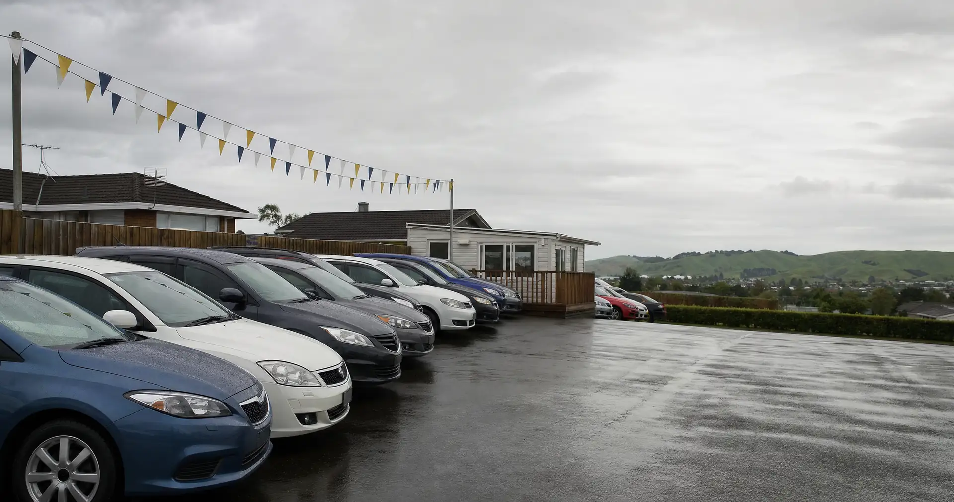 Rows of used cars on a wet suburban car yard under bunting on an overcast day